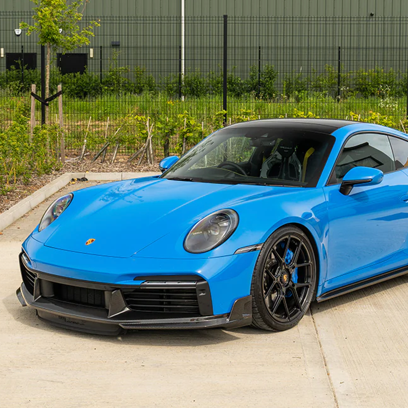 Blue Porsche sports car parked on a concrete surface with a green fence and plants in the background.